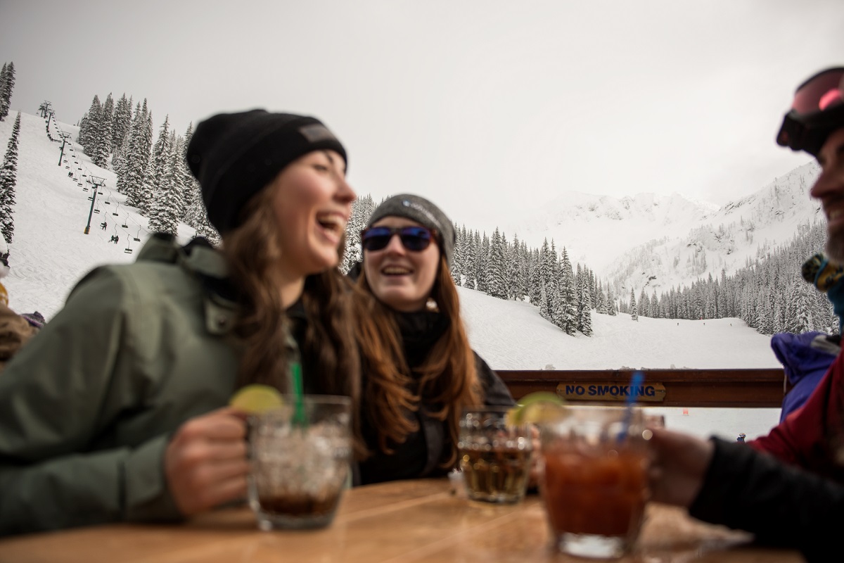 Three people sit on a patio drinking with the ski runs in the background