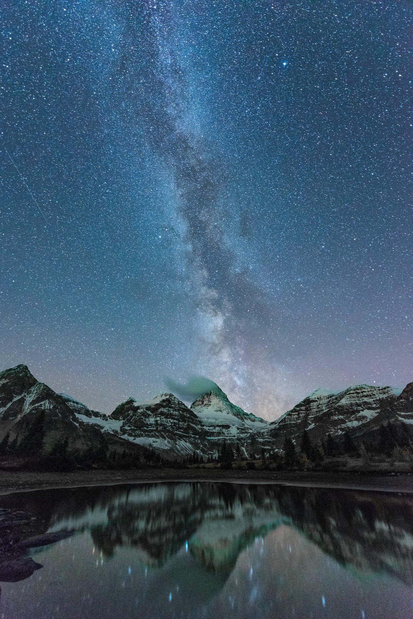 A clear alpine lake is in the foreground with snowy mountain peaks behind. Straight up from the peaks in the centre of the frame is the Milky Way in a star-filled sky.