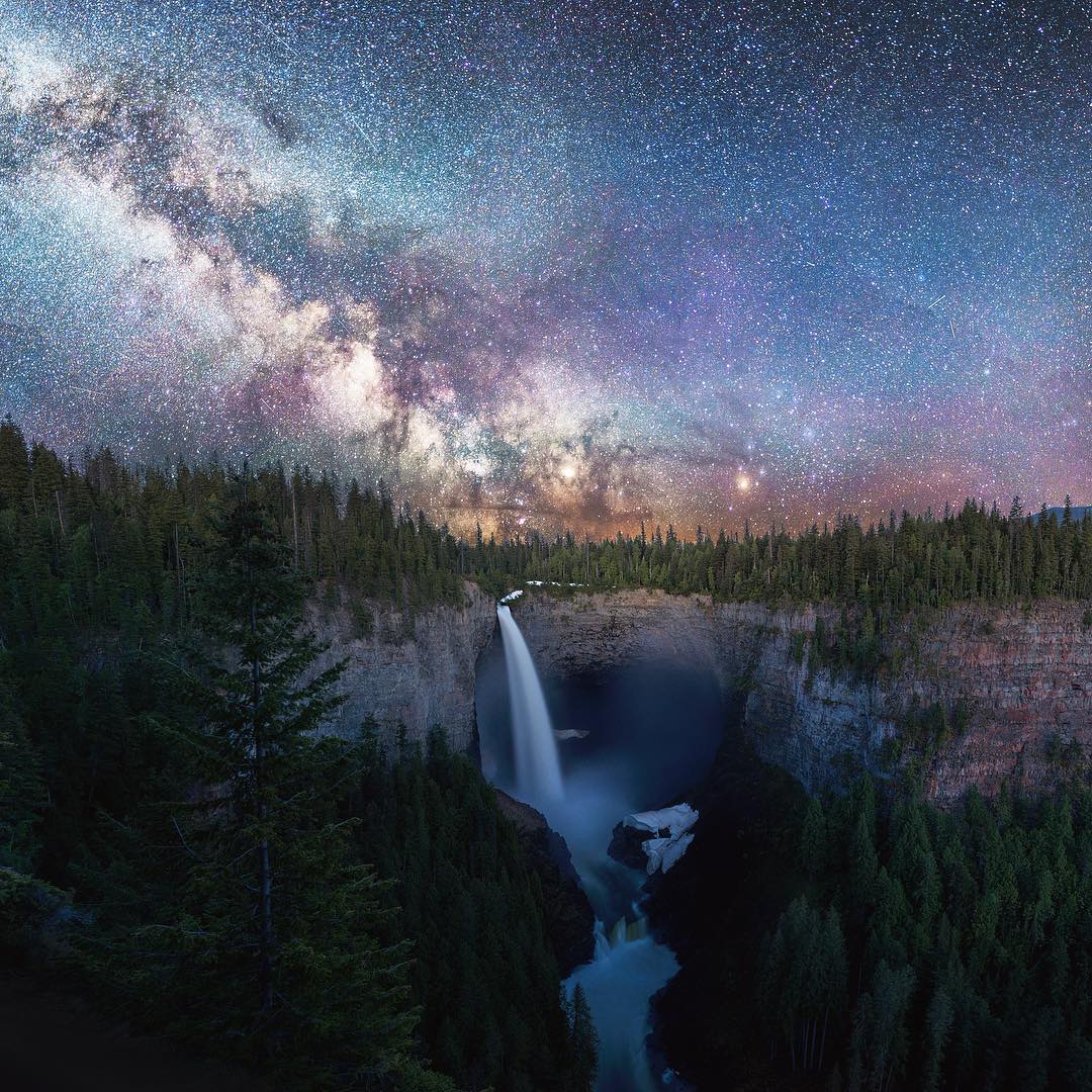 A steep waterfall plummets over a horseshoe-shaped cliff. There is thick forest all around and the incredibly clear night sky reveals a million stars and the Milky Way.