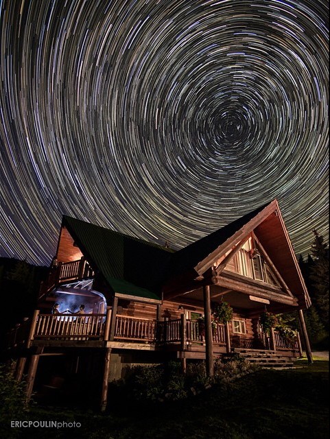 A wooden lodge sits in the centre of the frame and above it are circular streaks of light from a long exposure of the starry night sky