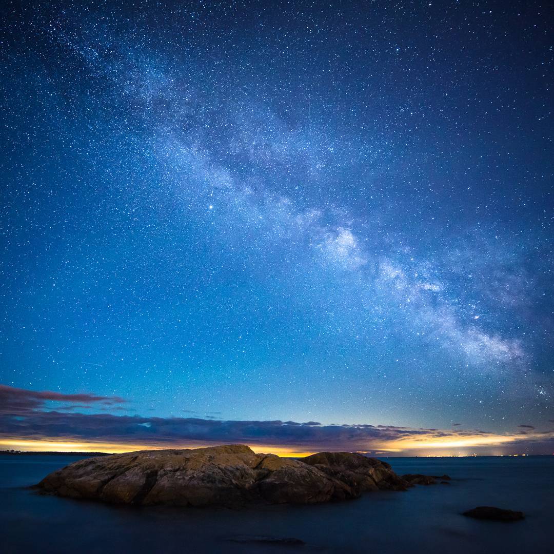 A small rocky islet sits just offshore. The last light from sunset is slightly visible on the horizon and there are a million stars in a clear blue sky. The Milky Way is visible.