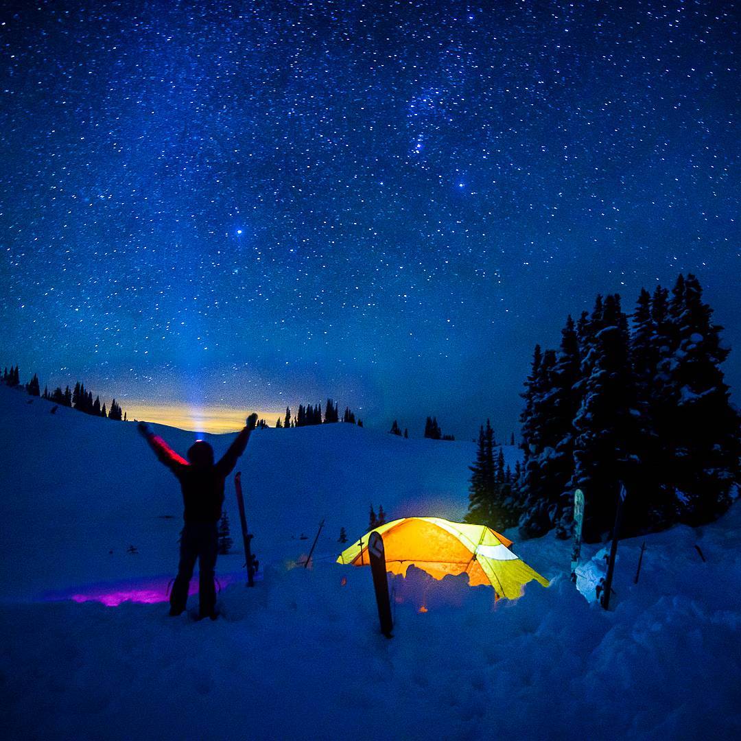 A figure stands in the snow, arms outstretched, beside a yellow tent lit from inside. The sky is midnight blue and filled with stars.