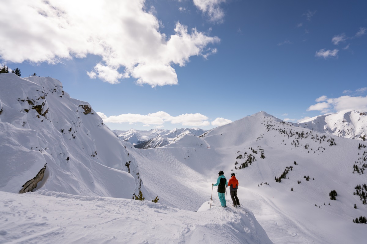 Two skiers in the alpine look out at the mountains on a blue sky day at Kicking Horse Mountain Resort