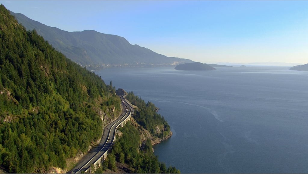 Aerial photo of the Sea-to-Sky Highway on a clear day. Cars move in both directions. The ocean is on one side and a steep mountain with trees is on the other. 