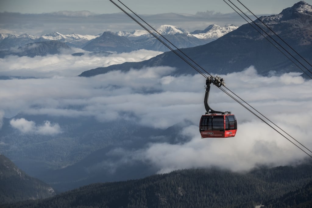 The red Peak 2 Peak Gondola moves along the wire. Clouds lift off the mountains below and snowcapped mountains are seen in the distance. 
