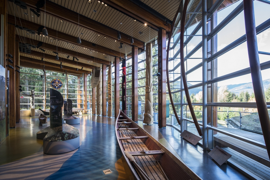 Inside the Squamish Lil'wat Cultural Centre. A canoe is beside large windows and other Indigenous signage and artwork is displayed. 