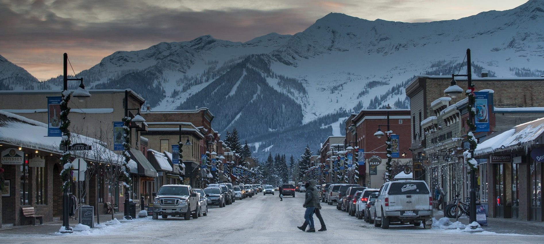 People walk through downtown Fernie in the snow. Snowcapped mountains frame the town in the distance.