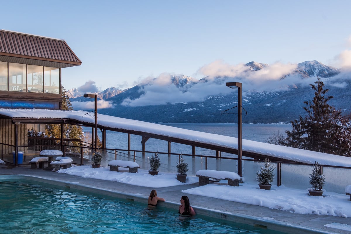 Two people soak in the Ainsworth Hot Springs, looking out at the lake. The deck is covered in snow. There are snow capped mountains in the distance.