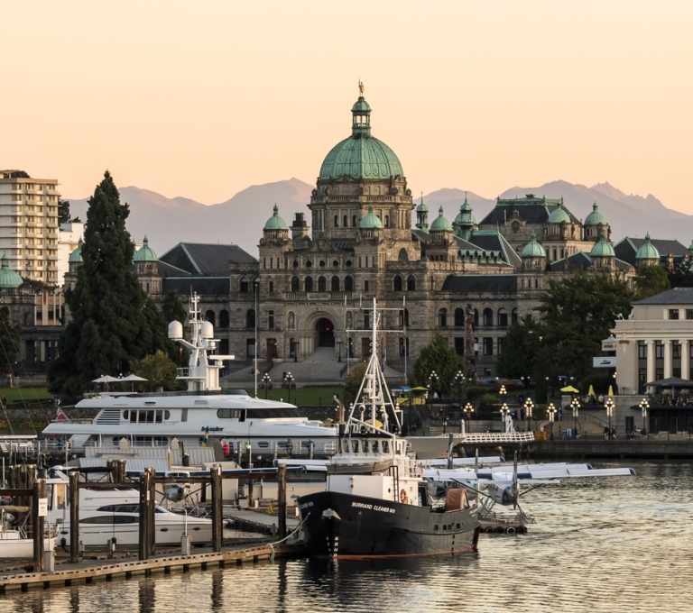 Downtown harbour in Victoria as the sunsets. A collection of boats are in the water, the Parliament buildings are seen in the background.