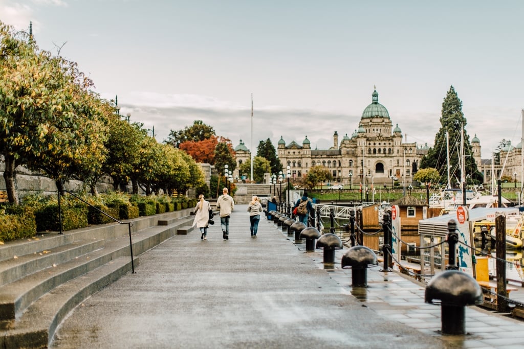 Three people walk along the pathway in the Inner Harbour on a cloudy day towards the Parliament buildings. 