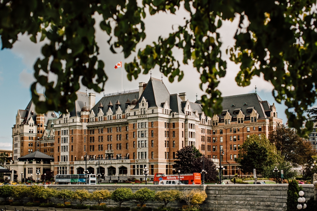 A side view of the Fairmont Empress hotel. Several tour busses are parked in front and the Canada flag flies from the top of the hotel. A tree is in the foreground of the camera lens