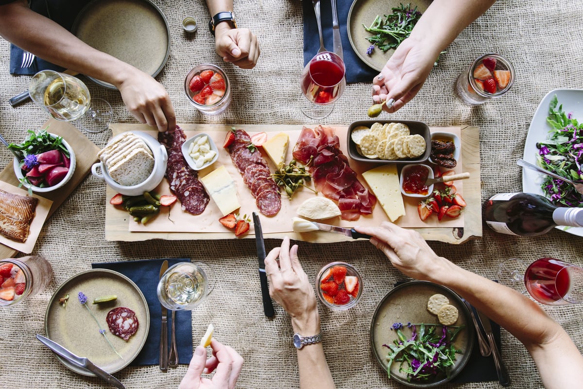 A top down shot of hands picking up food off a wooden charcuterie board with cheese, meets, jams and bread. There are four people dining total each with plates and glasses in front of them.