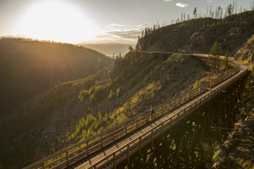 Two people look out into the distance from a trestle bridge on the Kettle Valley Rail Trail. The sun is setting and bright in the distance.