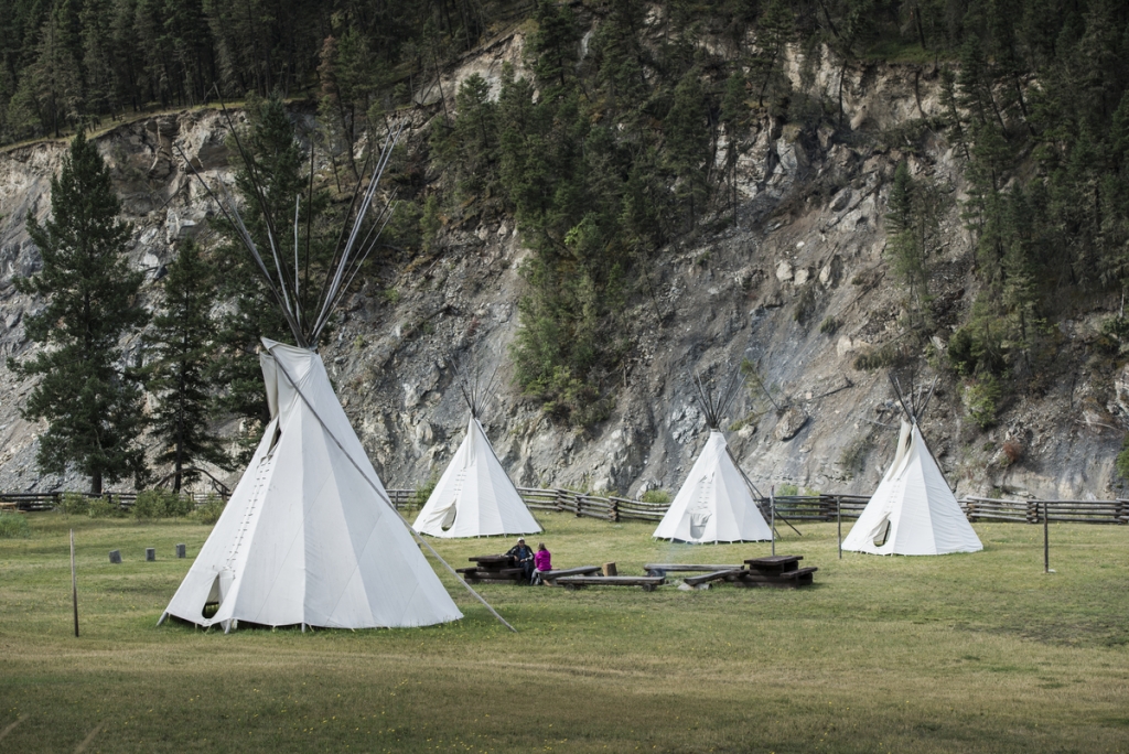 Four teepees at the Xat'sull Heritage Village. Two people sit together at a table in the middle. 