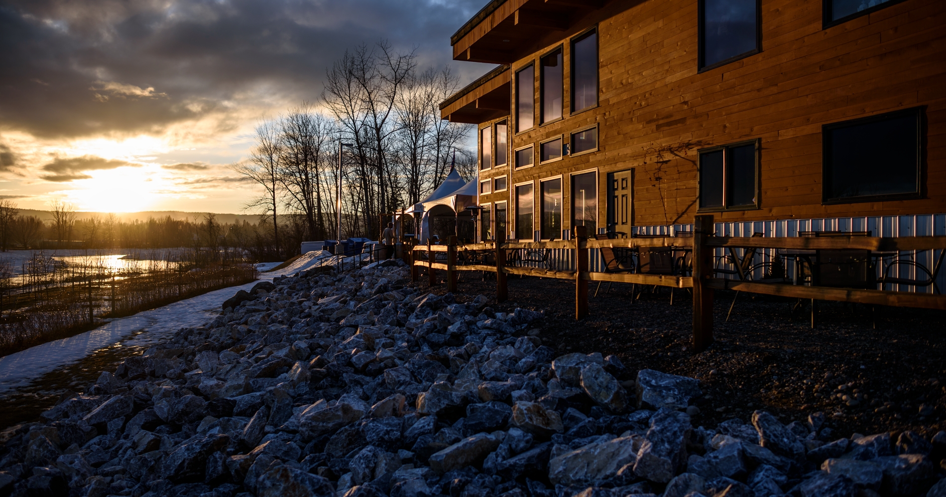 A wooden building sits on top of a rocky shore beside a river as the sun sets.