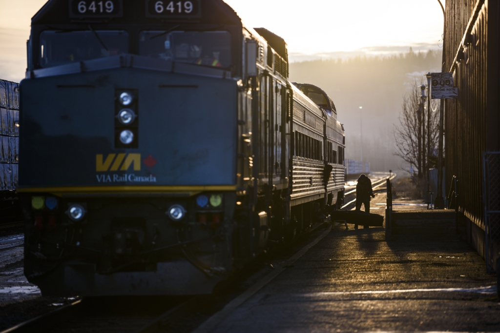 A close up of a VIA Rail train. The sun is setting behind the train. A person loads a bag onto the train at the station. 