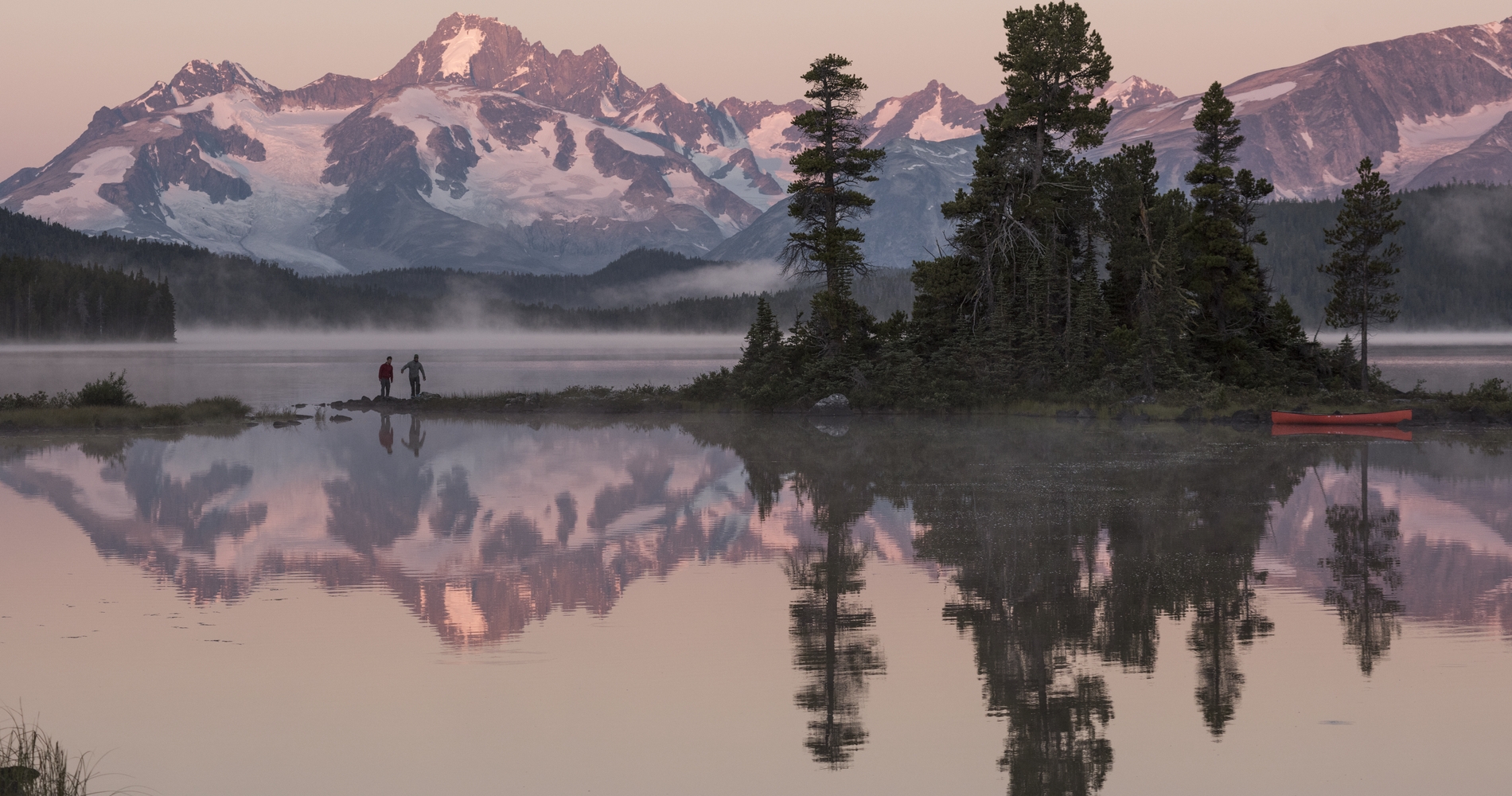 Two people walk along an island outcrop in a lake. The sun is setting on the mountains in the background. There is a stand of evergreen trees on the island and the image of the trees and mountains are reflected in the lake.