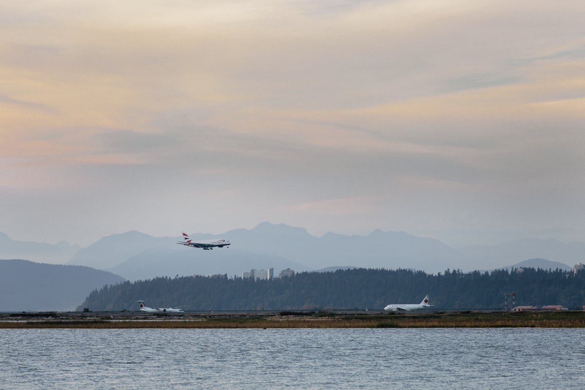 a plane lands at Vancouver International Airport. The photo is taken from across the Fraser River, and the North Shore Mountains are visible in the background.
