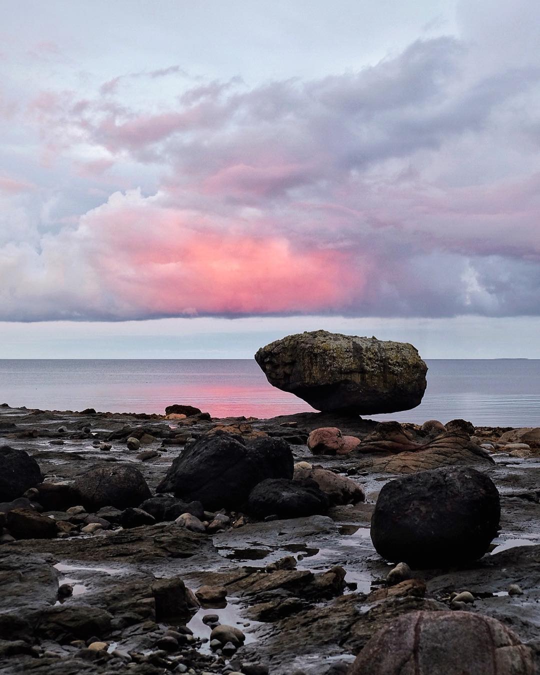 A large rock balances on a smaller rock on the beach at sunset. There are pink clouds in the sky.