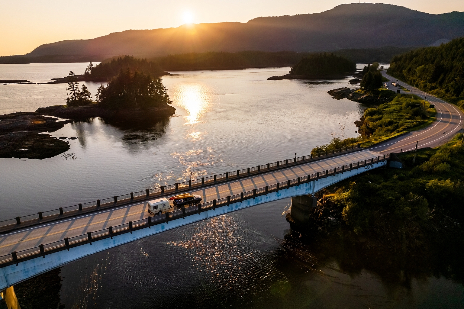 An truck with kayaks and a trailer drives across a bridge at sunset into Prince Rupert. Mountains are in the distance, and trees line the road past the bridge.