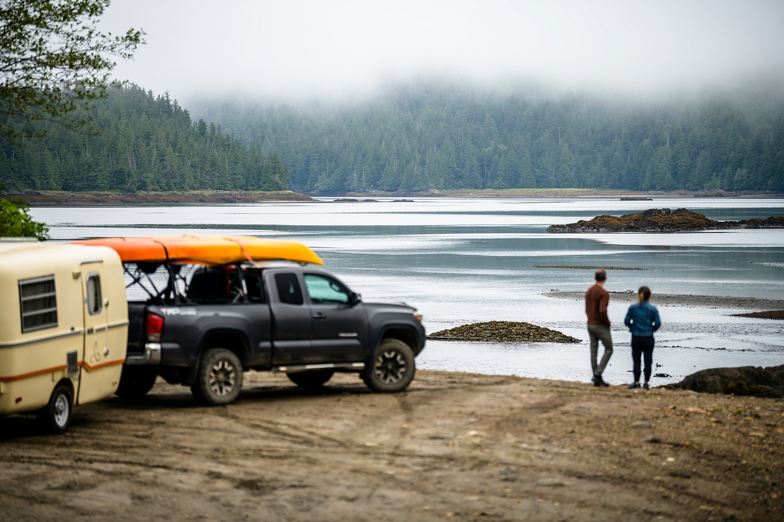 Two people stand at the shore looking out at the ocean. Fog covers the tops of the trees. A truck with two yellow kayaks pulling a trailer is parked on the beach.