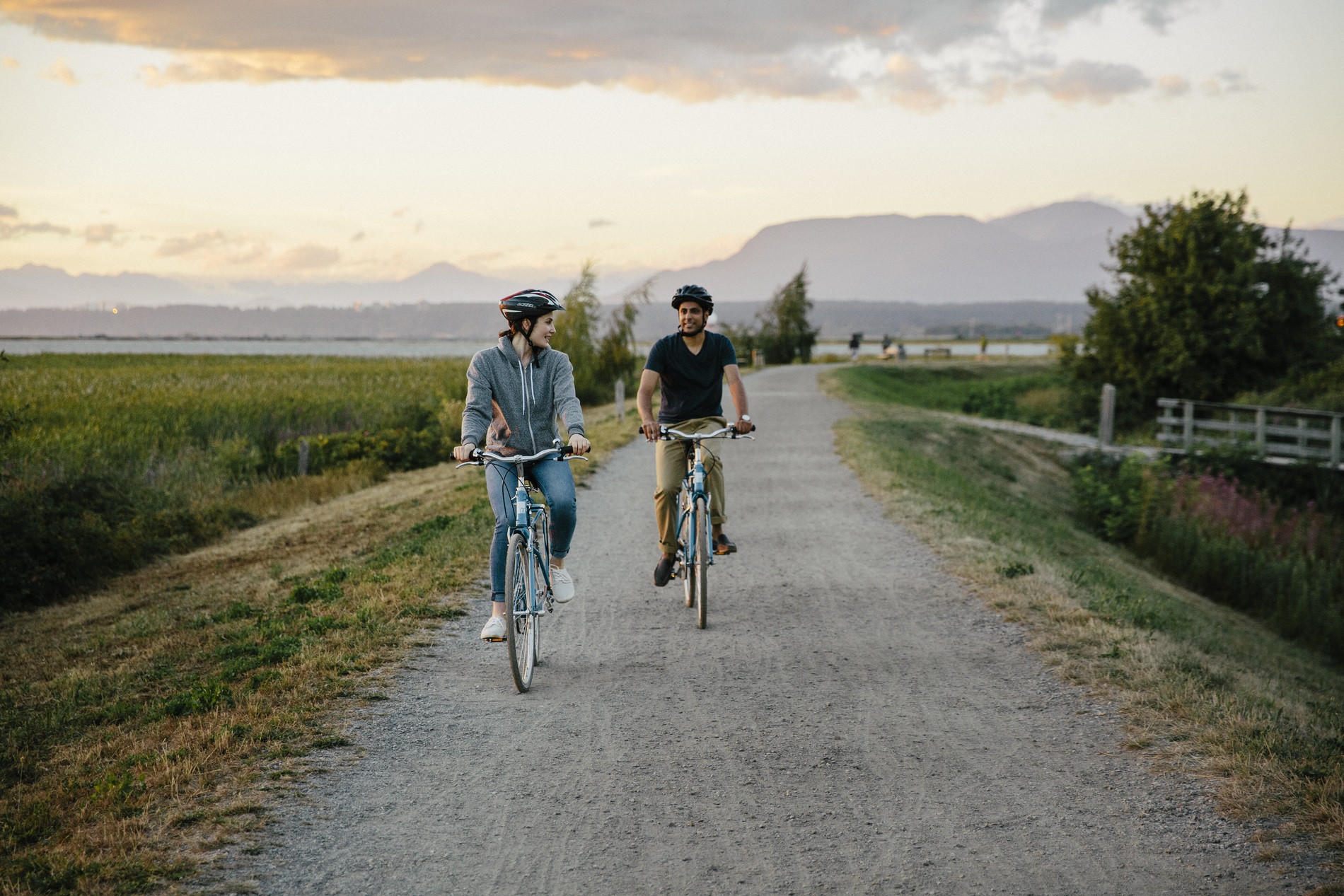 Two people ride bicycles down a flat gravel path. Fields and farmland are on both sides of the pathway.