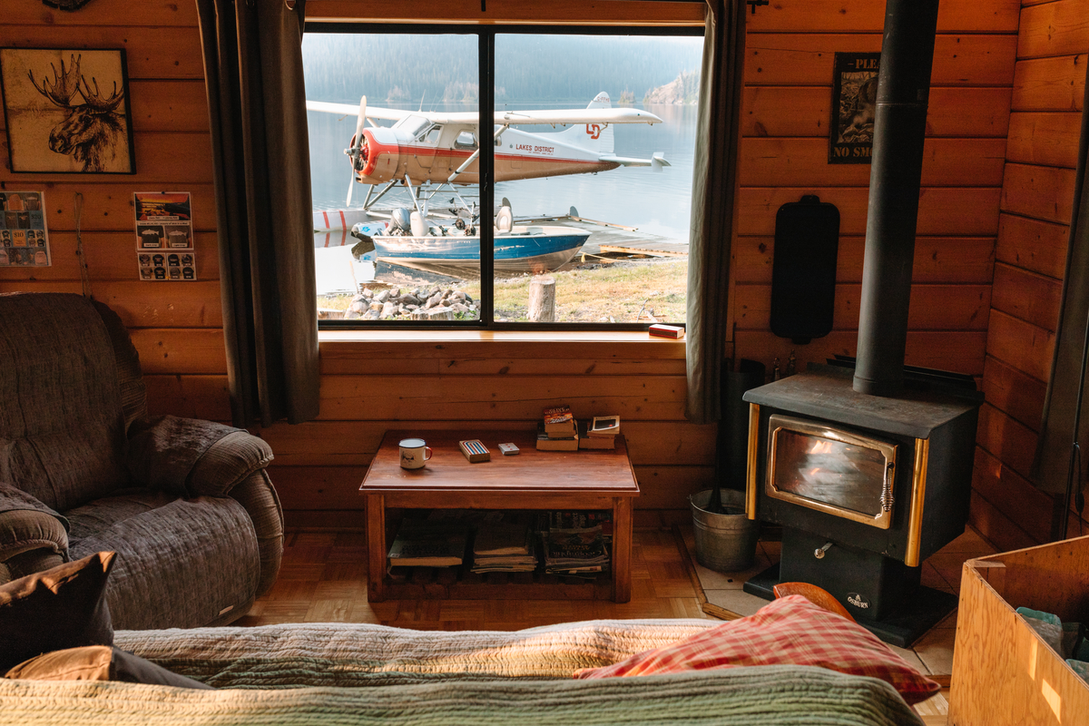 Interior log cabin view of Tesla Lake Lodge. The room has an armchair, small table, wood fireplace and sofa. The window looks out onto the beach. There is a small propeller plane and a boat on the beach.