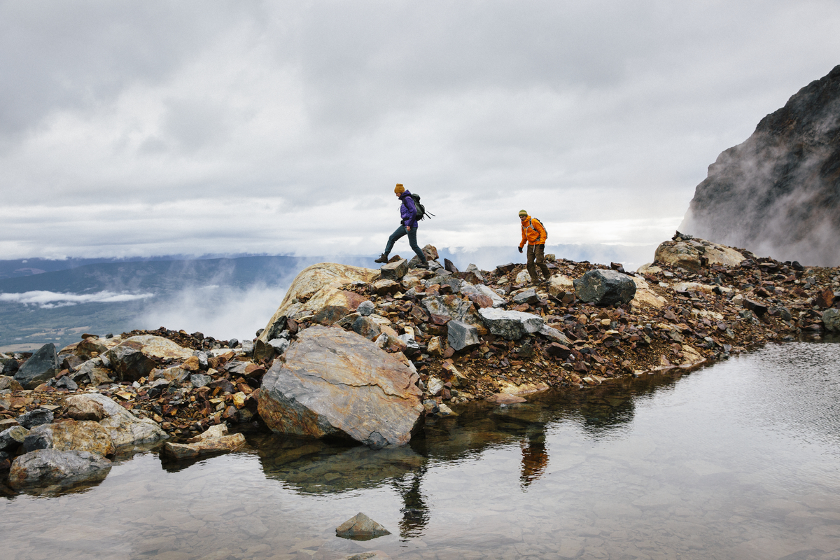 Two people walk along a rocky shoreline. One is wearing a blue jacket and pants, the other is in an orange jacket. There is water in the foreground and clouds overhead.