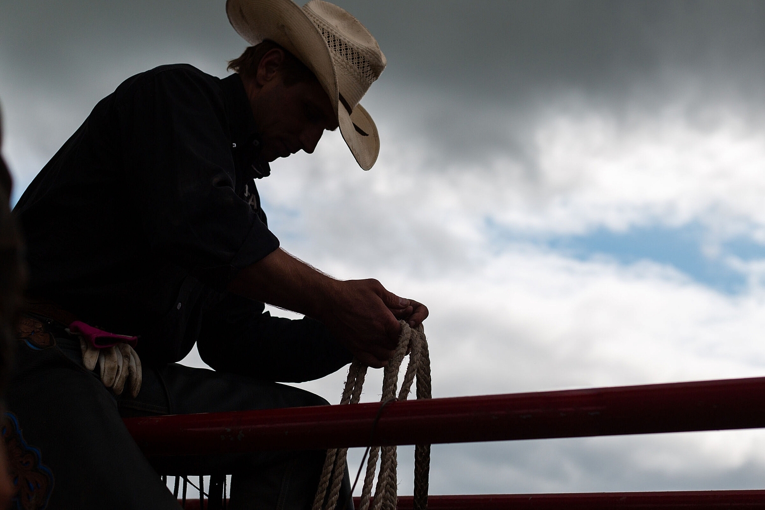 Silhouette of a cowboy in a hat sitting on a fence, holding a rope. Cloudy sky is in the background.