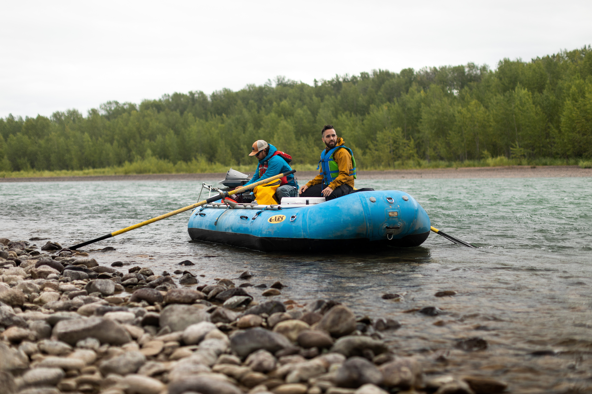 Two men sit in a blue raft on the Bulkley River. They are close to the rocky shore in the foreground. Green trees line the river bank on the opposite side.