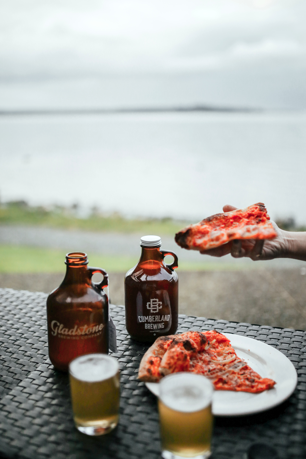 a patio table overlooking the ocean has two growlers, two glasses of beer, a plate with a slice of pizza, and from the right of the frame is a hand holding a second slice.