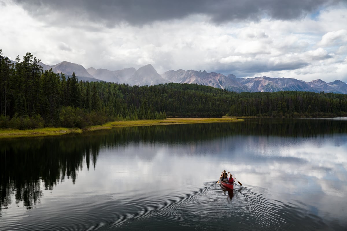 People paddle in a canoe across a lake surrounded by dense forest with mountains in the backdrop.