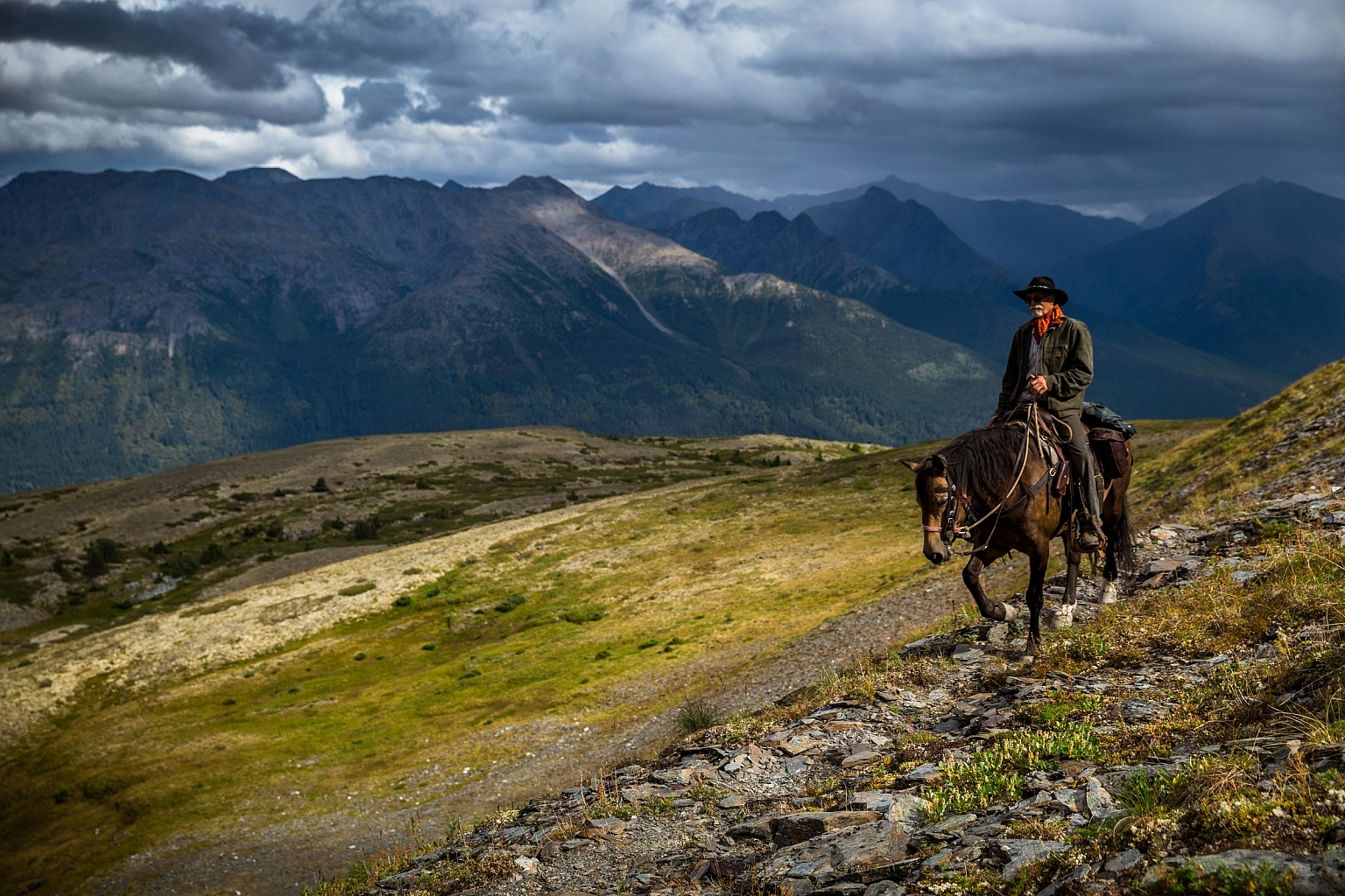 A lone horseback rider traverses the rugged countryside with rocks, sparse foliage and mountains as far as the eye can see.