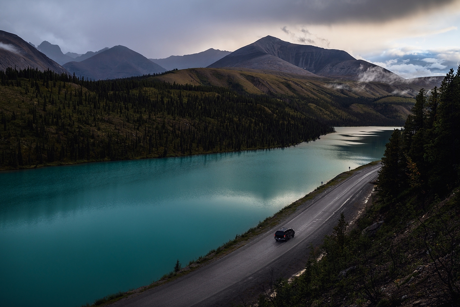 The Alaska Highway winds beside a turquoise lake with mountains in the background.