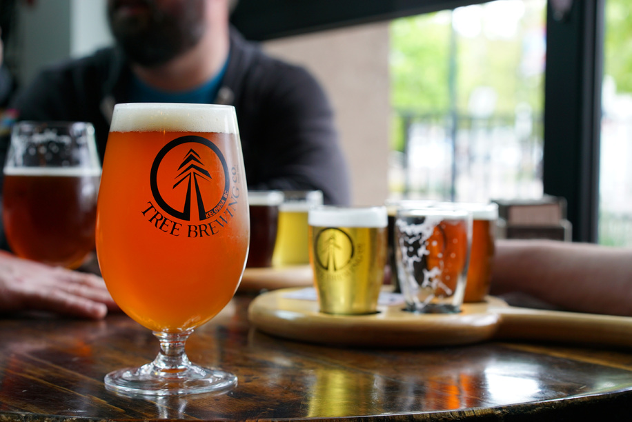 A close up of a beer in a glass at Tree Brewing. A person sits in a both in the background. Several small glasses of beer are arranged on the table.