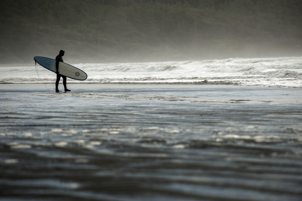 A person holds a surfboard and looks out at the water at Cox Bay