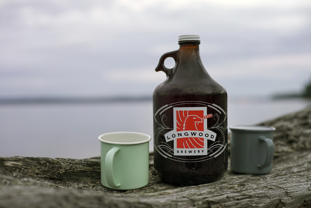 A growler from Longwood Brewery and a green mug sit o a log on the beach with the ocean behind.