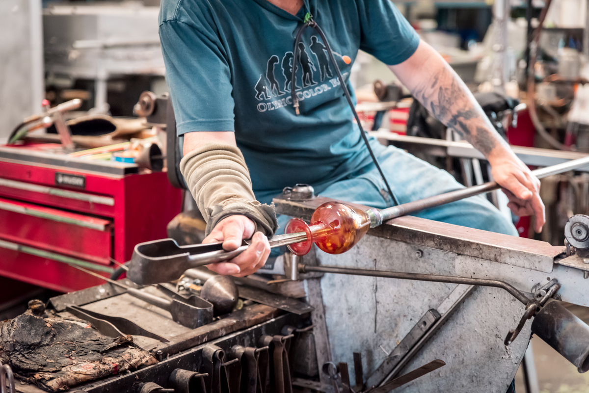 A close up of a person in a glass making workshop using metal tools to create a glass design.