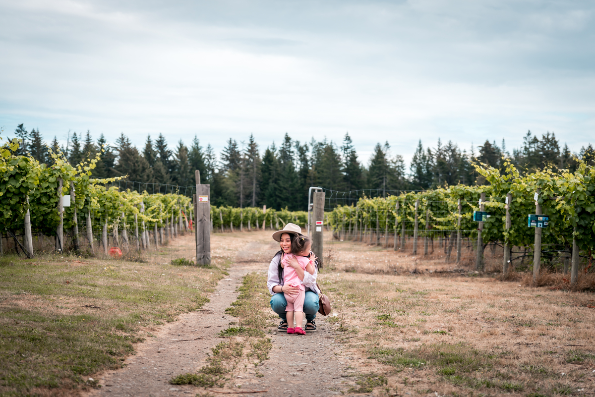 Yvonna Chow is embracing her young daughter in front of a vineyard. There is a fence and bushes behind them.