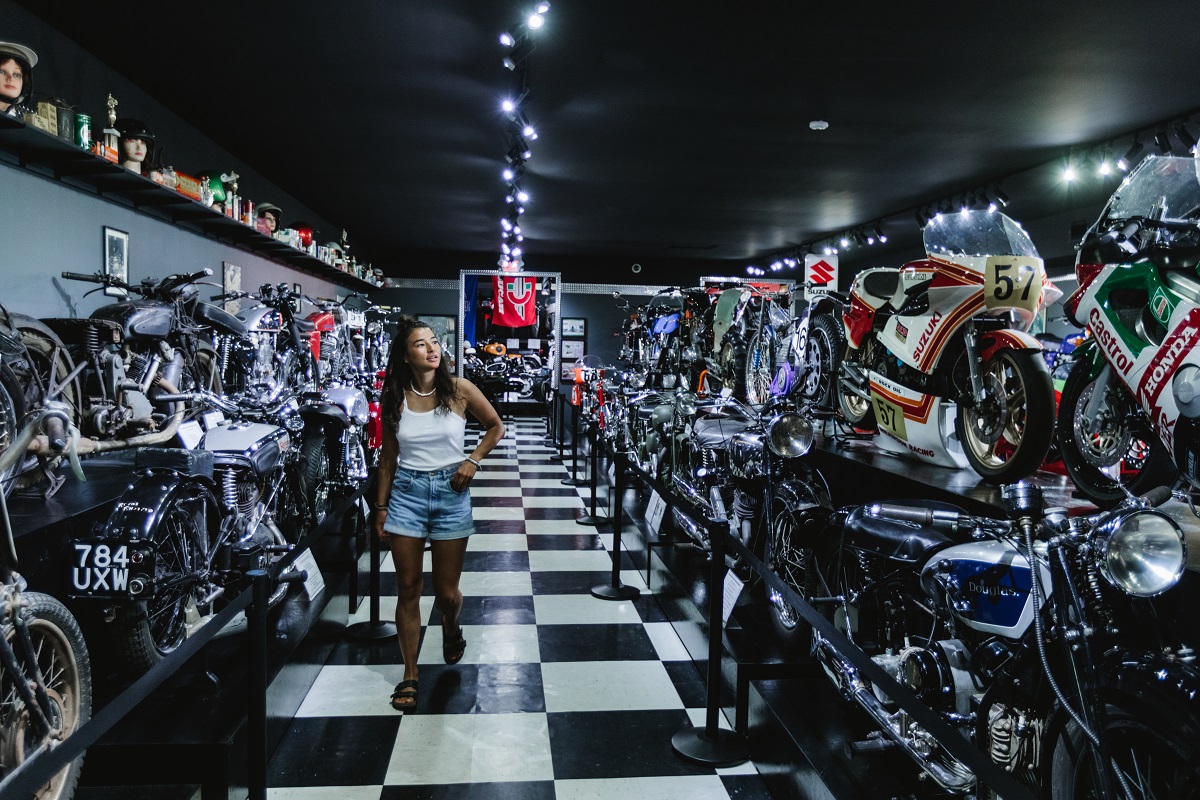 Mirae walks through the Sprokkets Cafe. There a numerous motorcycles on either side of her, and a black and white checkered floor.