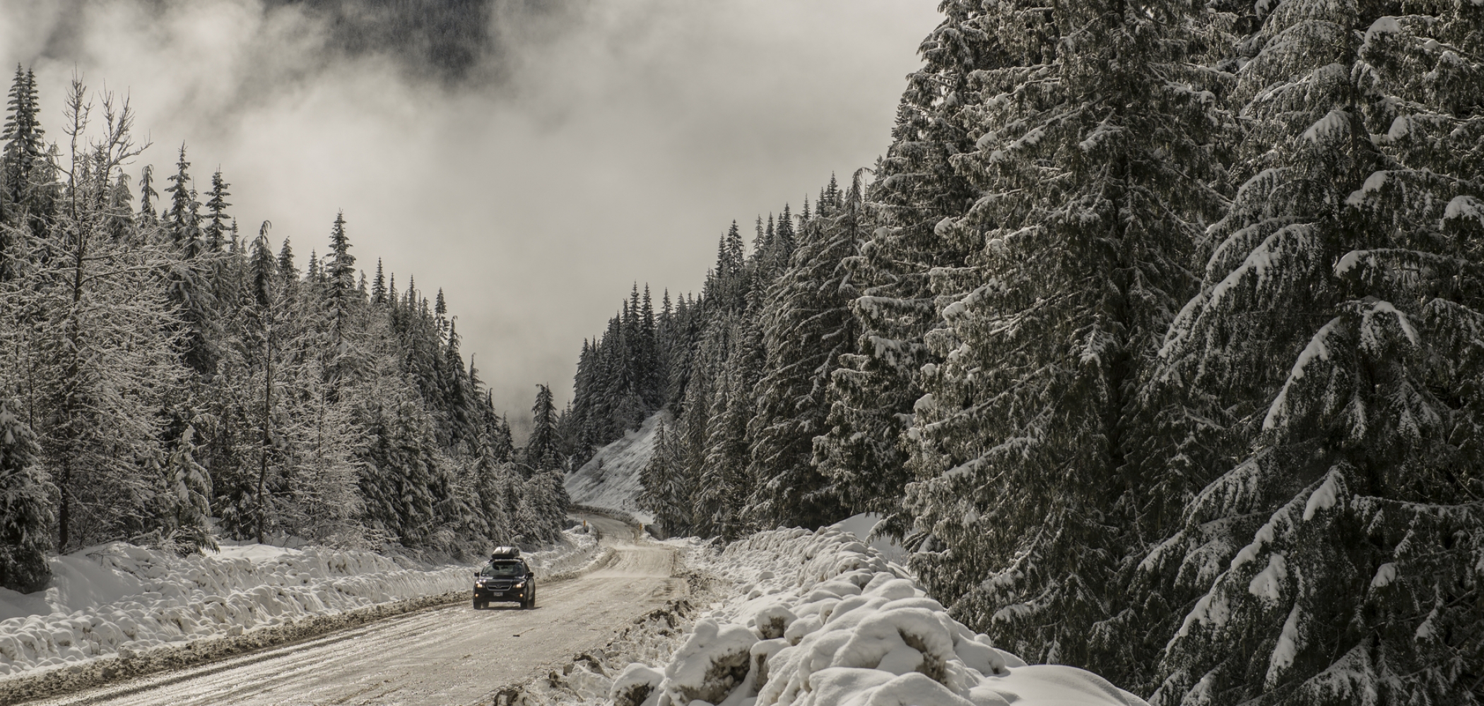 A car drives down an icy and snow covered road through the mountains with snow covered trees on each side.