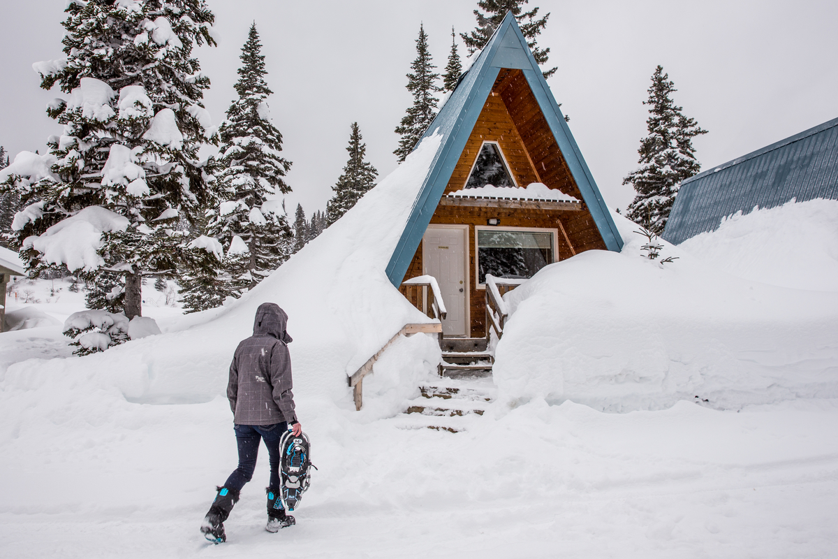 A person in a purple winter coat walks in the snow carrying snowshoes towards an A frame cabin framed by large snow banks