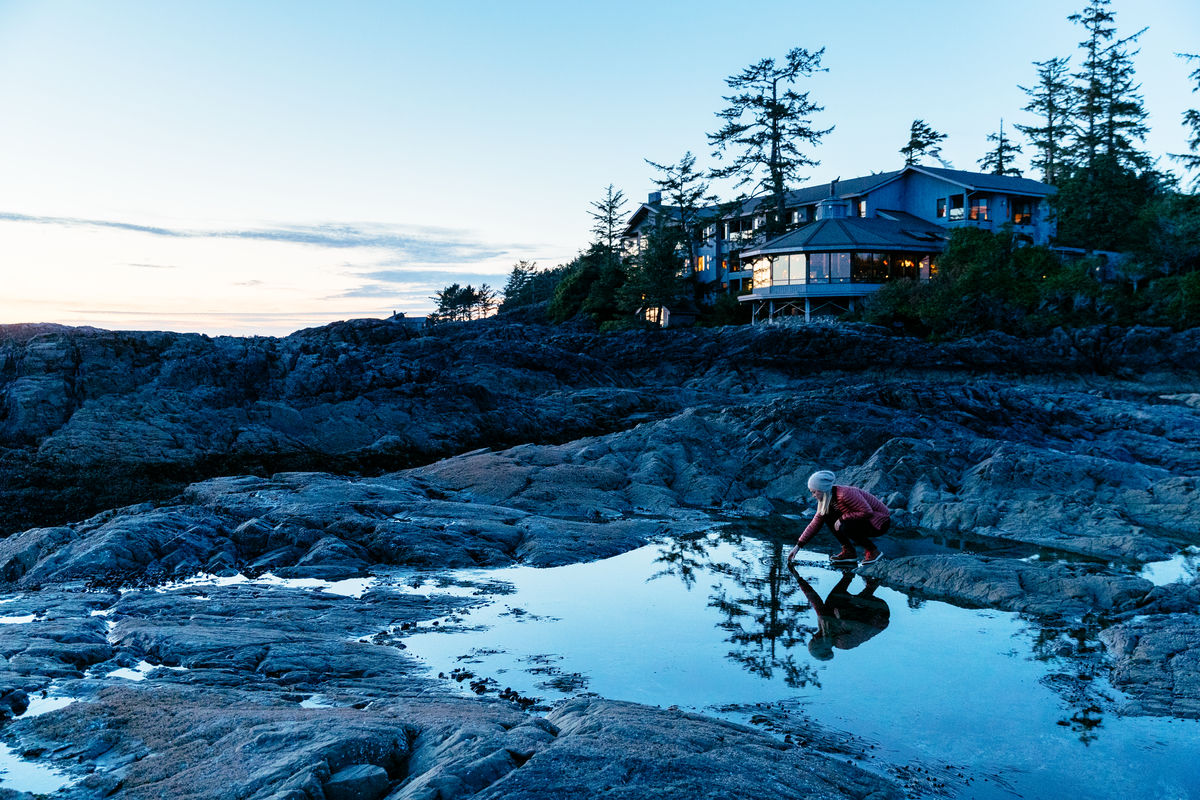 A women crouches down beside a rocky outcrop of water. The Wickaninnish Inn is behind her with the lights on.