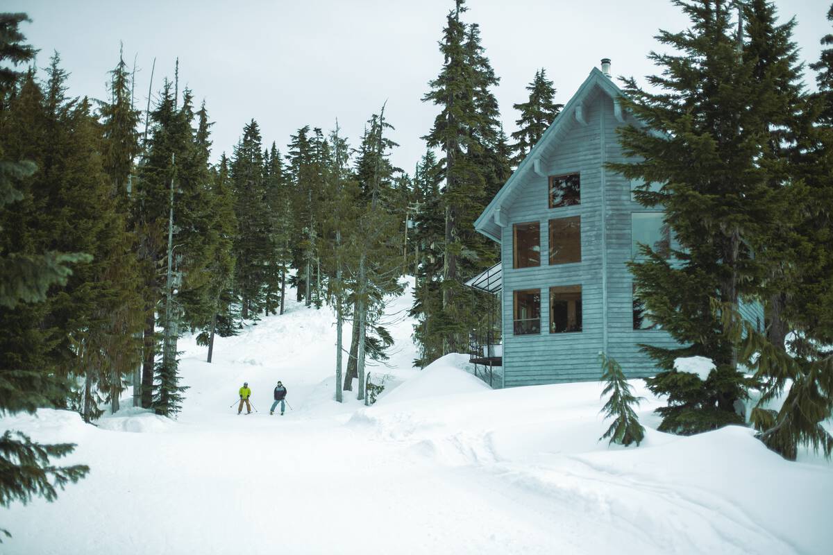 two people ski down a gentle run past a blue-coloured lodge on the right side of the frame