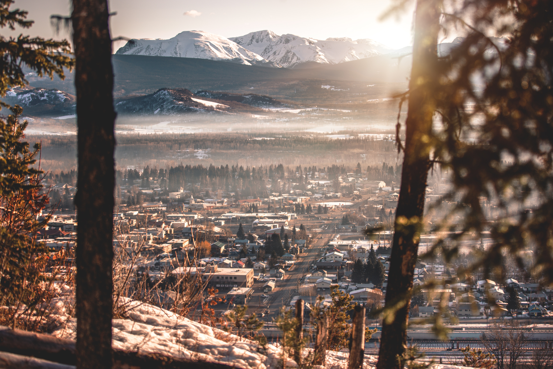 A look down at the town of Smithers in the winter from a high vantage point. The town is dusted in snow and there is a large mountain range in the background.