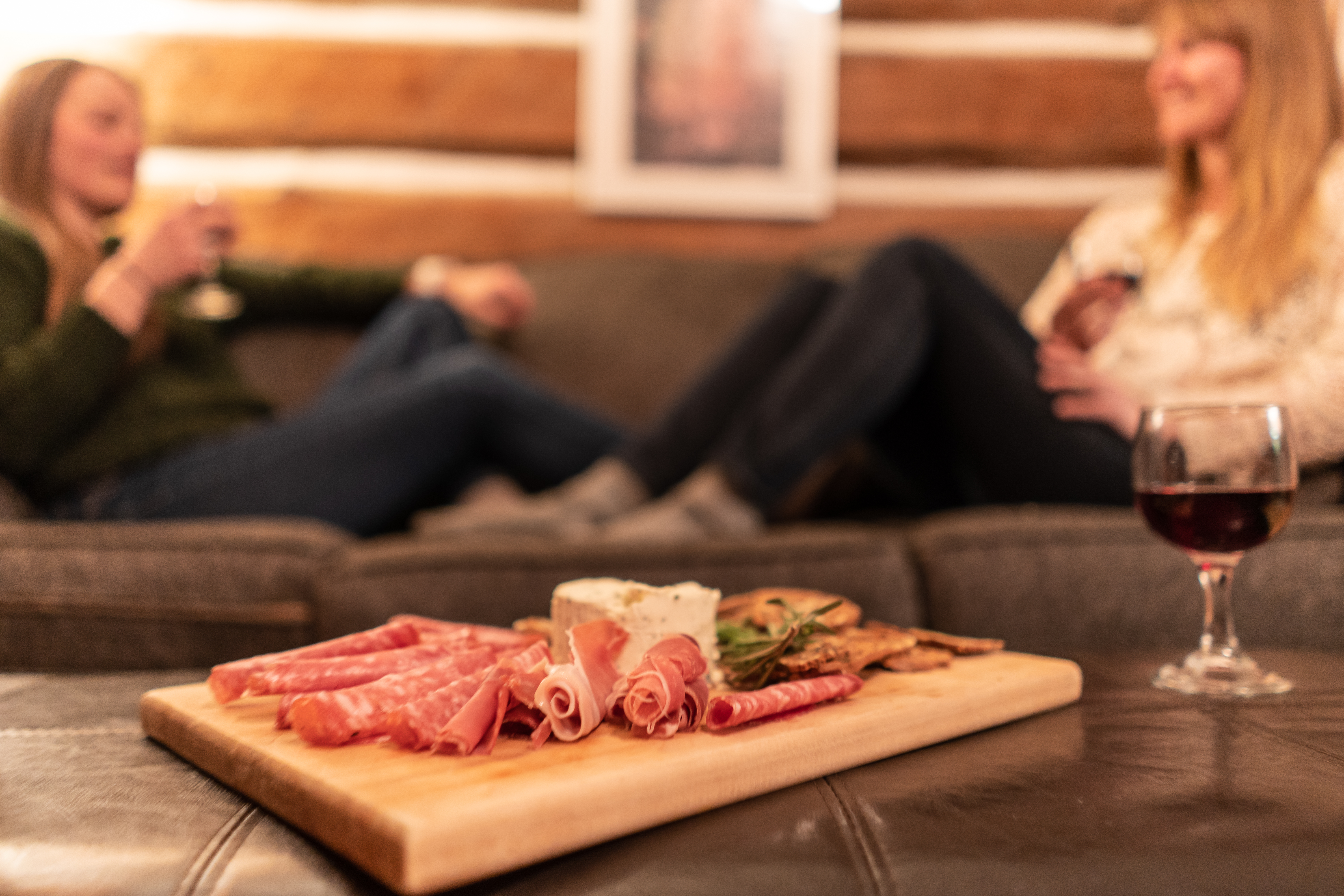 A charcuterie board with meat and cheese is on a table in the foreground, two women sit on a couch in the background.m