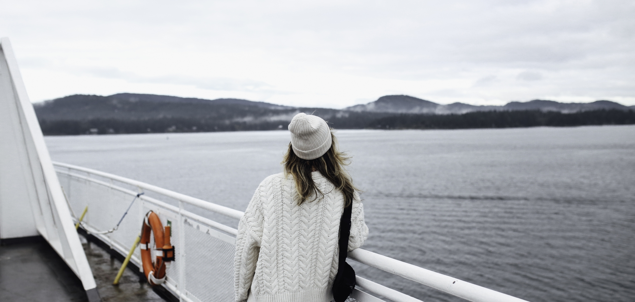 Person with a white sweater and matching toque leans against a railing on a BC Ferries vessel through the Gulf Islands toward Vancouver Island