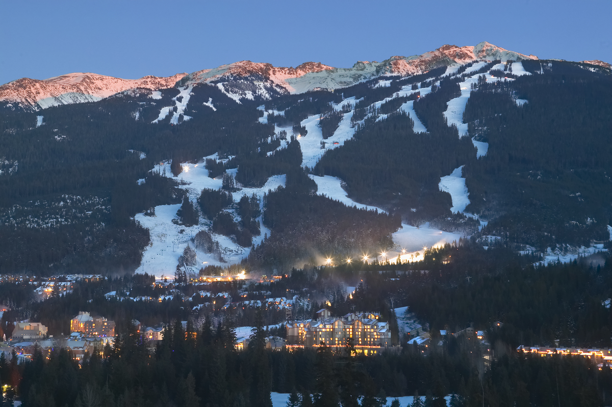 A nighttime view of Whistler resort in the wintertime. There are lights at the base of the mountain, and the village in the distance.