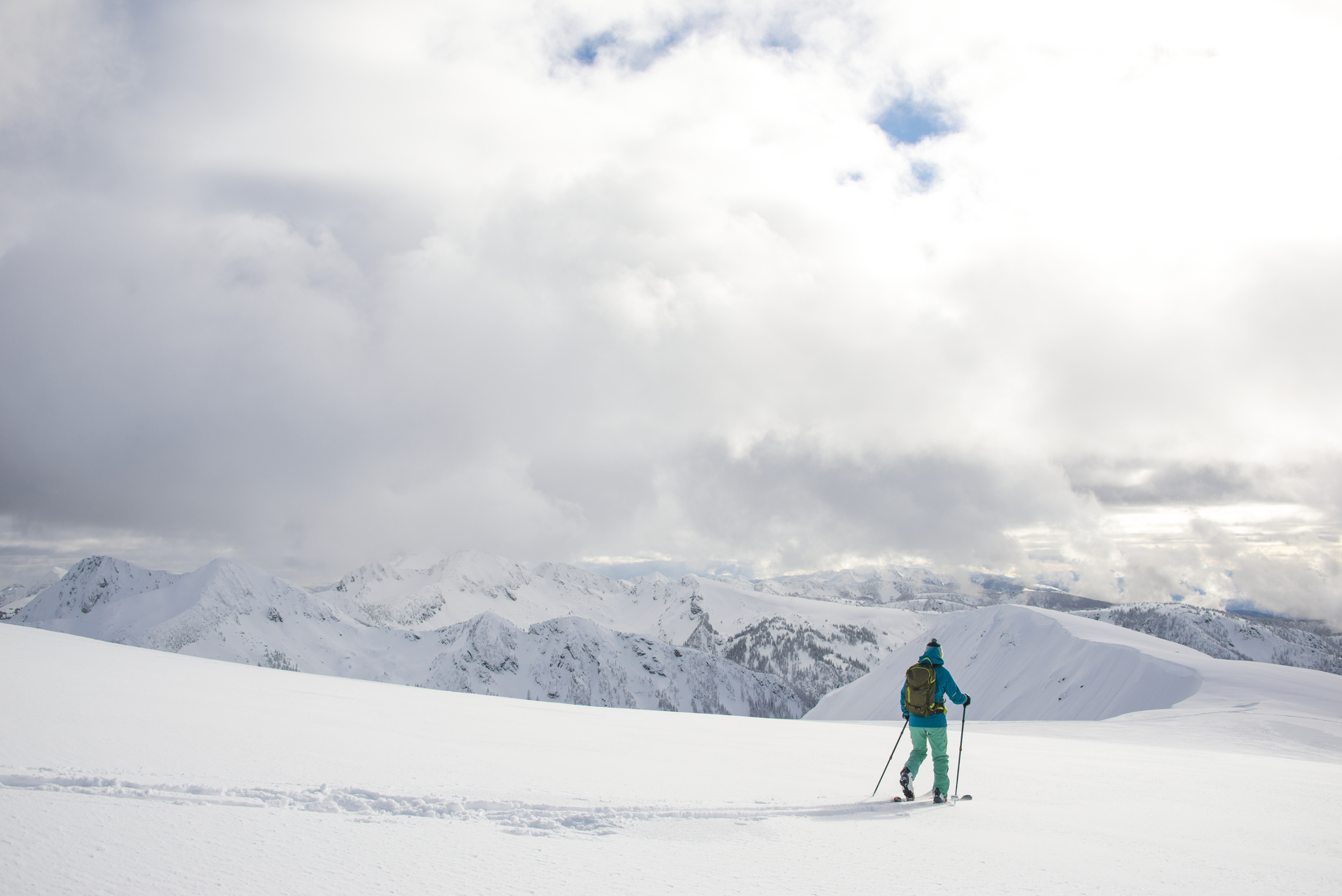 Backcountry skiing in the Valkyr Range of the southern Selkirk Mountains. The skier is wearing green snow pants and a blue jacket moving away from the camera. Clouds obscure the sky.