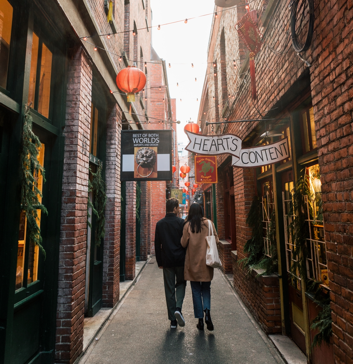 Couple wander through the narrow Fan Tan Alley in Victoria walking away from the camera. Strings of lights and lanterns are overhead.